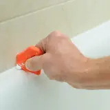 Mano usa spatola arancione per rimuovere silicone da bordo vasca bianca, vicino a piastrelle beige.