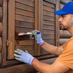 Uomo con guanti blu e cappellino blu restaura scuri in legno. Scopri i prezzi per il restauro scuri in legno.