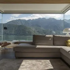 Soggiorno moderno con vista panoramica su lago e montagne, reso possibile da ampi infissi di ultima generazione. Divano, poltrona e tappeto creano un'atmosfera accogliente.
