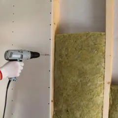 Installazione di pannelli isolanti per un maggiore comfort acustico. Una mano con guanto bianco avvita un pannello di cartongesso su una struttura in legno con isolamento.