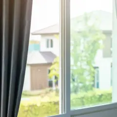 Tende grigie aperte su una finestra, lasciando intravedere case e alberi verdi. Si percepisce una leggera corrente d'aria in casa.