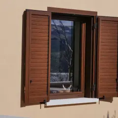 Facciate con persiane marroni che incorniciano una finestra aperta, riflettendo un paesaggio alberato.