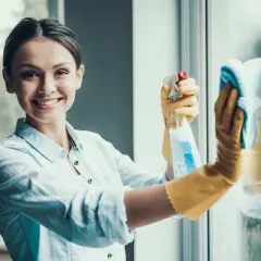 Donna sorridente con guanti gialli pulisce finestre in alluminio con spray e panno. Manutenzione serramenti in alluminio.