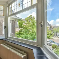 Vista da una finestra con vetri colorati, che si apre a destra o sinistra, su un quartiere alberato con auto parcheggiate.