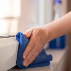Mano pulisce davanzale con panno blu. Un flacone di detergente è sullo sfondo. Scopri come pulire gli scuri in legno.