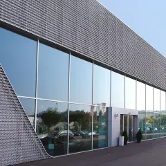 Moderne facciate architettoniche con ampie vetrate riflettono il cielo e gli alberi. La struttura presenta un rivestimento metallico perforato con un design angolare distintivo.