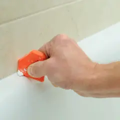 Mano usa spatola arancione per rimuovere silicone da bordo vasca bianca, vicino a piastrelle beige.