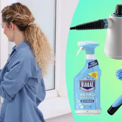 Donna pulisce vetri esterni molto sporchi con spray e panno. Accanto, prodotti per la pulizia: pulitore a vapore, spray Viakal e lavavetri.
