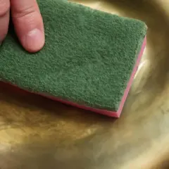 Preparazione per verniciare infissi alluminio dorato: una mano lucida una superficie dorata con una spugna abrasiva verde.