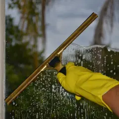 Mano con guanto giallo usa un tergivetro per pulire i vetri delle finestre, lasciando una scia di schiuma e gocce.