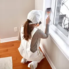 Donna con tuta da lavoro e cappellino bianco mentre è intenta a verniciare infissi in alluminio con un pennello.