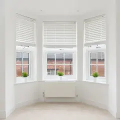 Angolo luminoso con tre finestre inglesine, piante verdi e un radiatore bianco.