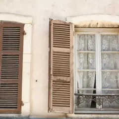 Due finestre d'epoca con persiane in legno marrone. Una è chiusa, l'altra è aperta rivelando tende di pizzo bianco.
