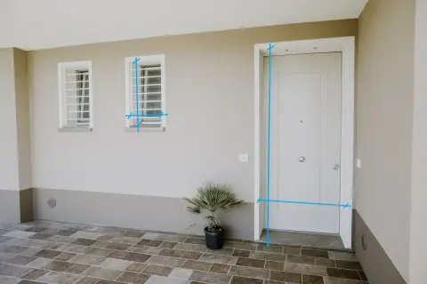 Misurazioni sul vano finestra e sulla porta d'ingresso. Due finestre con inferriate e una porta bianca su un muro beige.