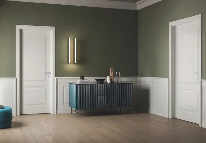Interno con pareti verde salvia e boiserie bianca, porte bianche, un mobile blu e un pouf turchese.