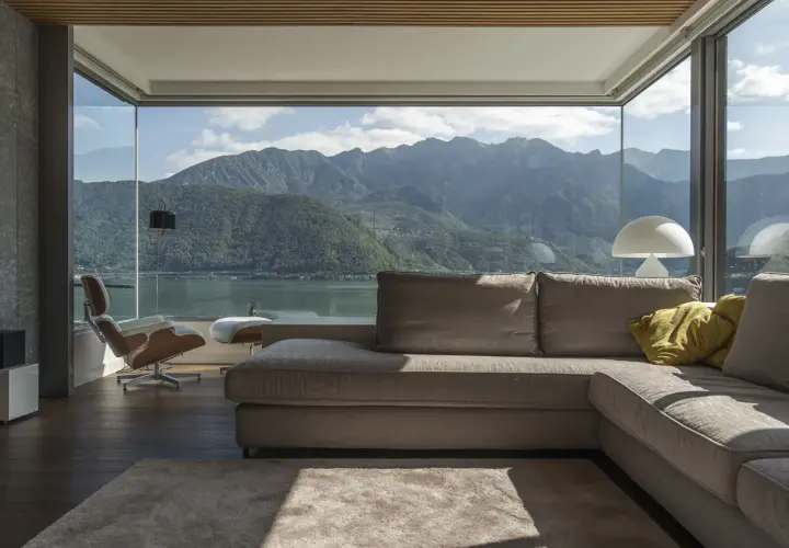 Soggiorno moderno con vista panoramica su lago e montagne, reso possibile da ampi infissi di ultima generazione. Divano, poltrona e tappeto creano un'atmosfera accogliente.