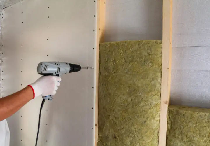Installazione di pannelli isolanti per un maggiore comfort acustico. Una mano con guanto bianco avvita un pannello di cartongesso su una struttura in legno con isolamento.