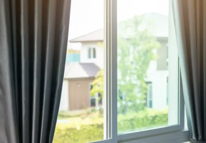 Tende grigie aperte su una finestra, lasciando intravedere case e alberi verdi. Si percepisce una leggera corrente d'aria in casa.