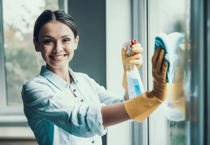 Donna sorridente con guanti gialli pulisce finestre in alluminio con spray e panno. Manutenzione serramenti in alluminio.