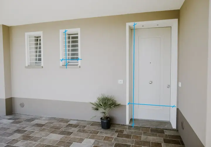Misurazioni sul vano finestra e sulla porta d'ingresso. Due finestre con inferriate e una porta bianca su un muro beige.