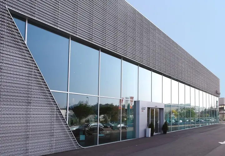 Moderne facciate architettoniche con ampie vetrate riflettono il cielo e gli alberi. La struttura presenta un rivestimento metallico perforato con un design angolare distintivo.