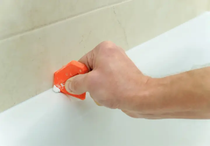 Mano usa spatola arancione per rimuovere silicone da bordo vasca bianca, vicino a piastrelle beige.