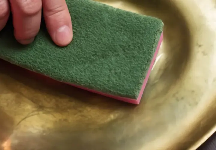 Preparazione per verniciare infissi alluminio dorato: una mano lucida una superficie dorata con una spugna abrasiva verde.