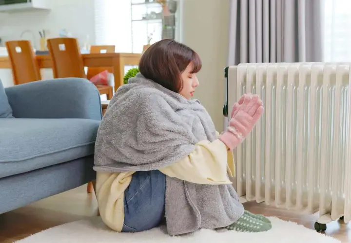 Ragazza avvolta in una coperta, con guanti rosa, vicino a un termosifone acceso, ma la casa è fredda nonostante i termosifoni.