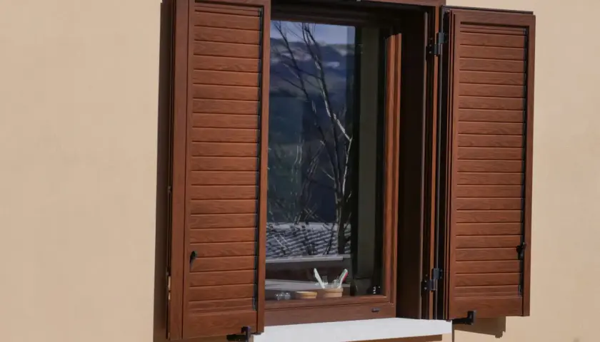Facciate con persiane marroni che incorniciano una finestra aperta, riflettendo un paesaggio alberato.