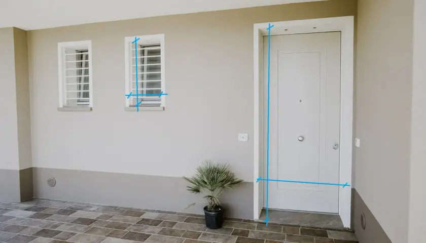 Misurazioni sul vano finestra e sulla porta d'ingresso. Due finestre con inferriate e una porta bianca su un muro beige.