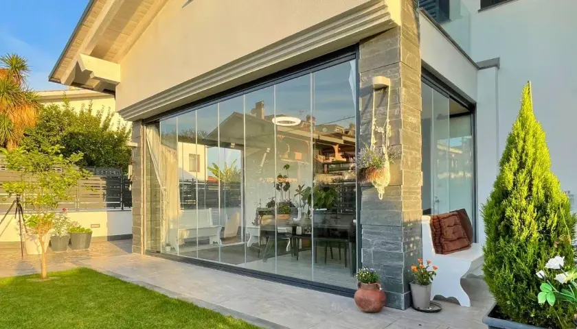 Moderna chiusura veranda in vetro, con arredi interni visibili. Un'elegante soluzione che rispetta la chiusura veranda normativa, valorizzando lo spazio esterno.