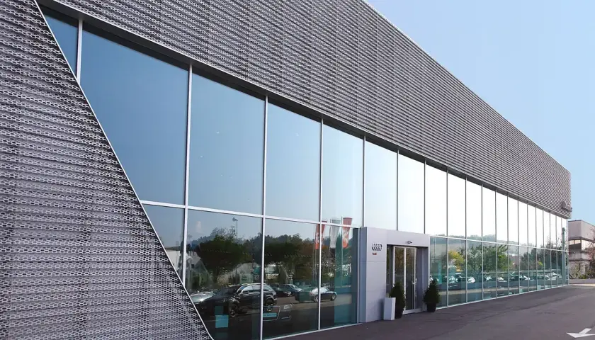 Moderne facciate architettoniche con ampie vetrate riflettono il cielo e gli alberi. La struttura presenta un rivestimento metallico perforato con un design angolare distintivo.