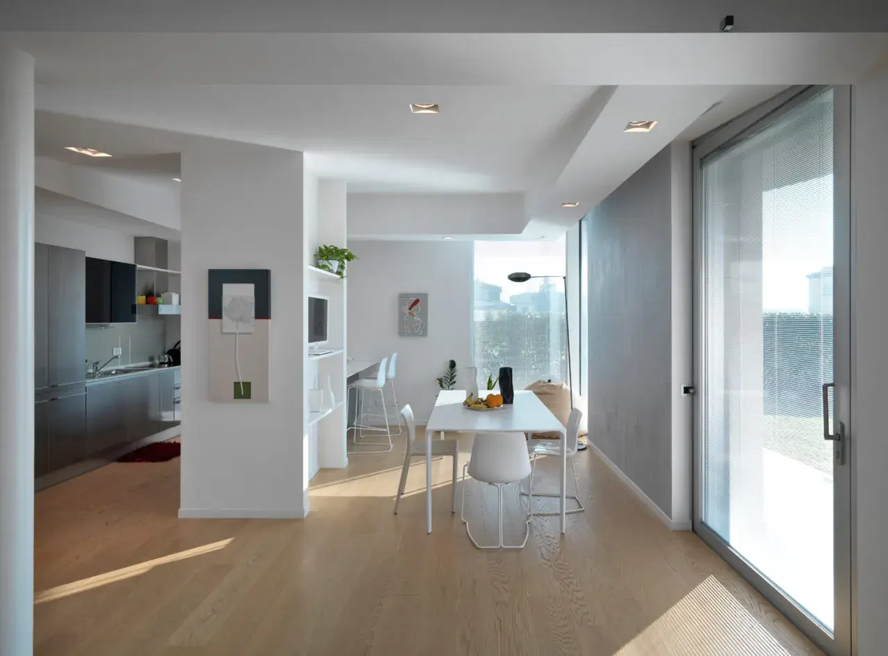 Interno moderno con cucina a vista, zona pranzo e un'ampia vetrata che sembra allargare una finestra in condominio, inondando di luce naturale.