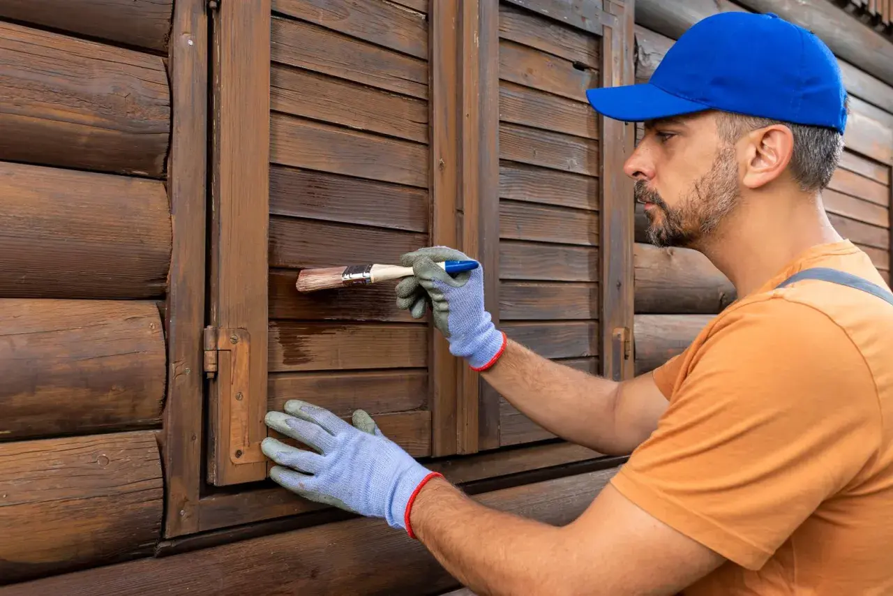 Uomo con guanti blu e cappellino blu restaura scuri in legno. Scopri i prezzi per il restauro scuri in legno.