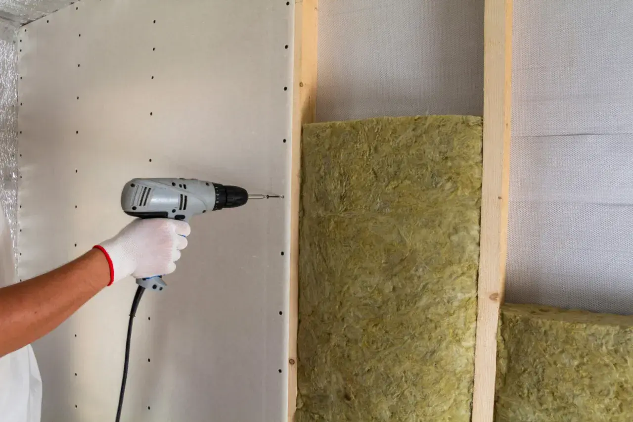 Installazione di pannelli isolanti per un maggiore comfort acustico. Una mano con guanto bianco avvita un pannello di cartongesso su una struttura in legno con isolamento.
