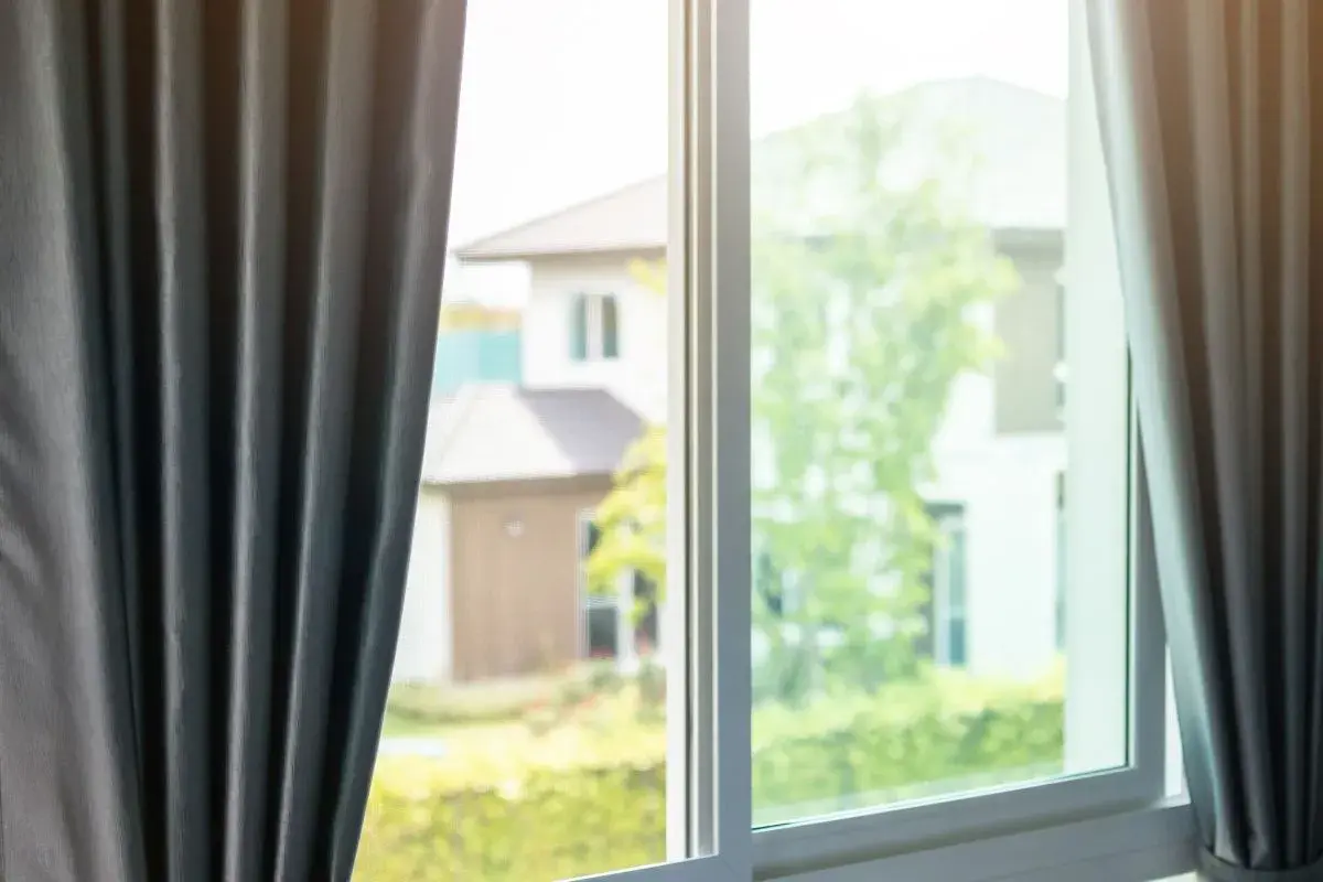 Tende grigie aperte su una finestra, lasciando intravedere case e alberi verdi. Si percepisce una leggera corrente d'aria in casa.