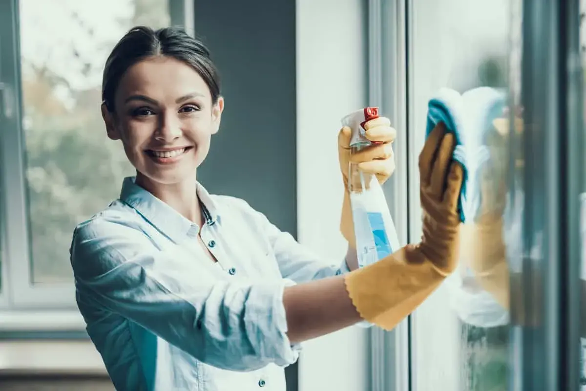 Donna sorridente con guanti gialli pulisce finestre in alluminio con spray e panno. Manutenzione serramenti in alluminio.
