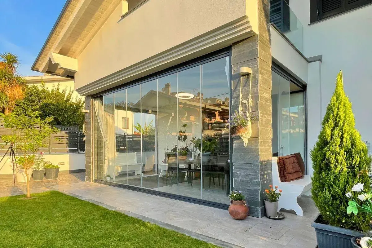 Moderna chiusura veranda in vetro, con arredi interni visibili. Un'elegante soluzione che rispetta la chiusura veranda normativa, valorizzando lo spazio esterno.