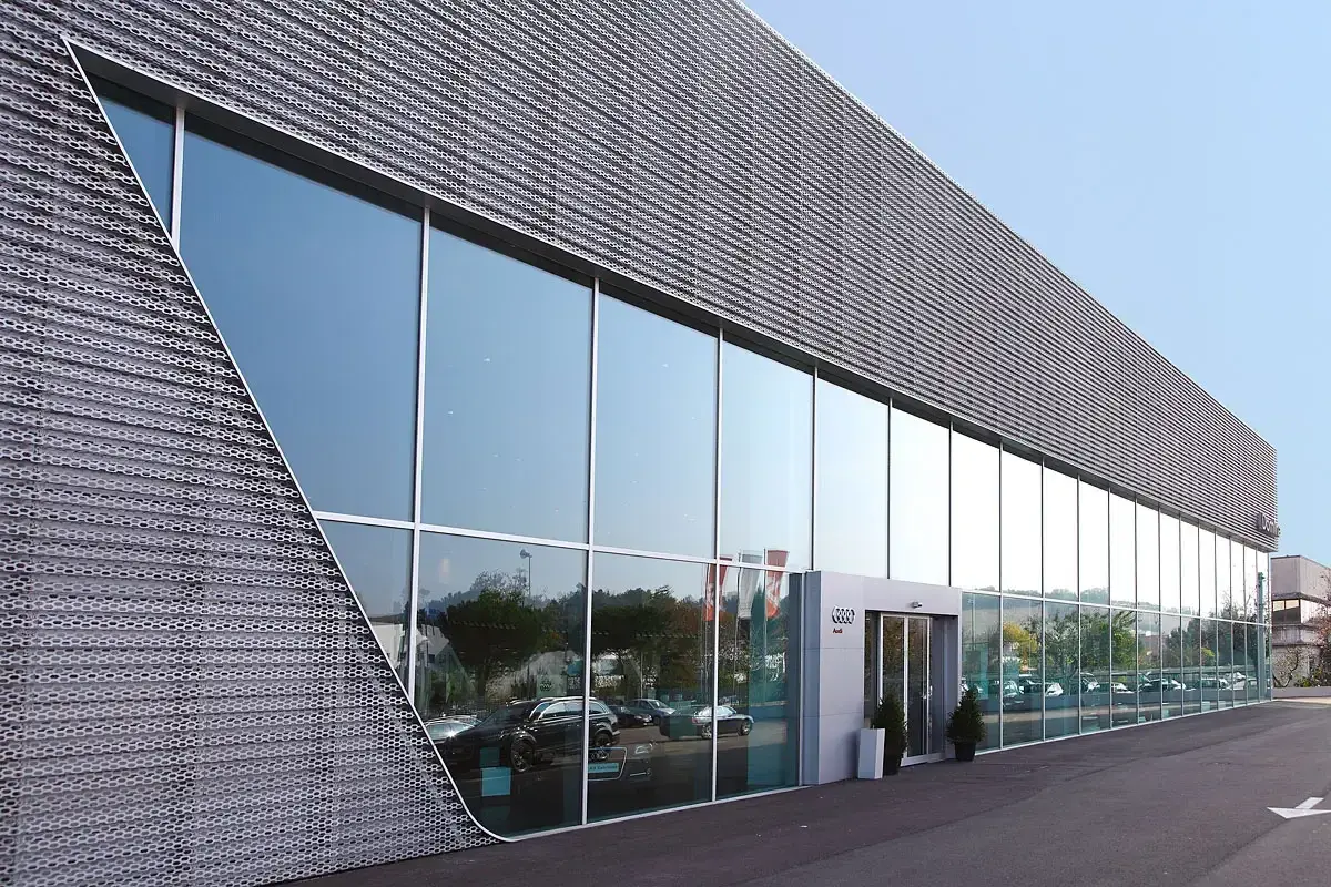 Moderne facciate architettoniche con ampie vetrate riflettono il cielo e gli alberi. La struttura presenta un rivestimento metallico perforato con un design angolare distintivo.