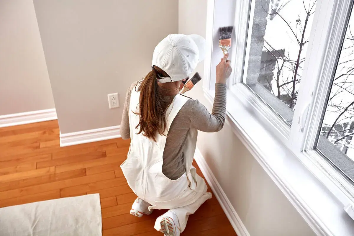 Donna con tuta da lavoro e cappellino bianco mentre è intenta a verniciare infissi in alluminio con un pennello.