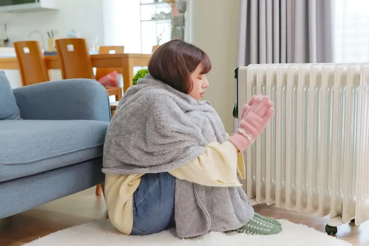 Ragazza avvolta in una coperta, con guanti rosa, vicino a un termosifone acceso, ma la casa è fredda nonostante i termosifoni.