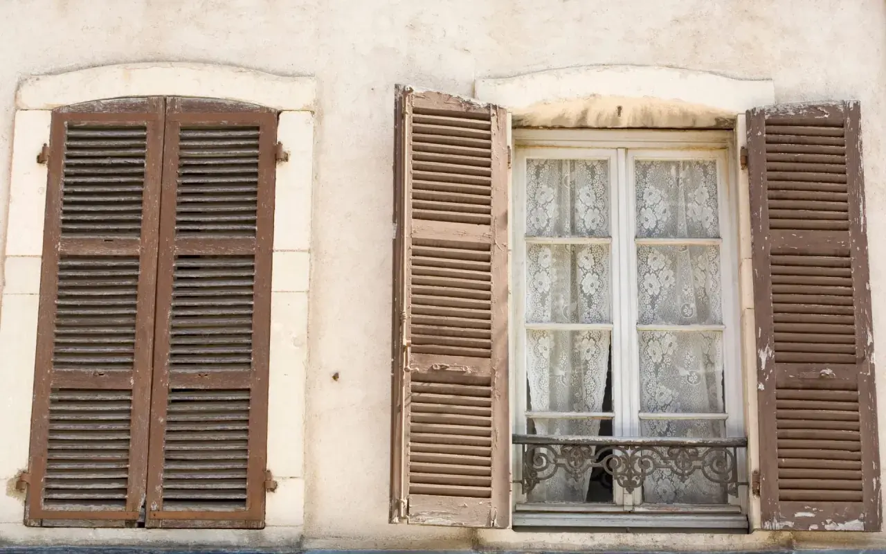 Due finestre d'epoca con persiane in legno marrone. Una è chiusa, l'altra è aperta rivelando tende di pizzo bianco.