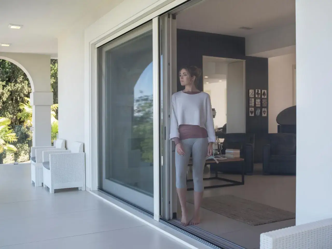 Ragazza guarda fuori da una moderna finestra senza telaio, godendosi la vista del patio con sedie bianche.