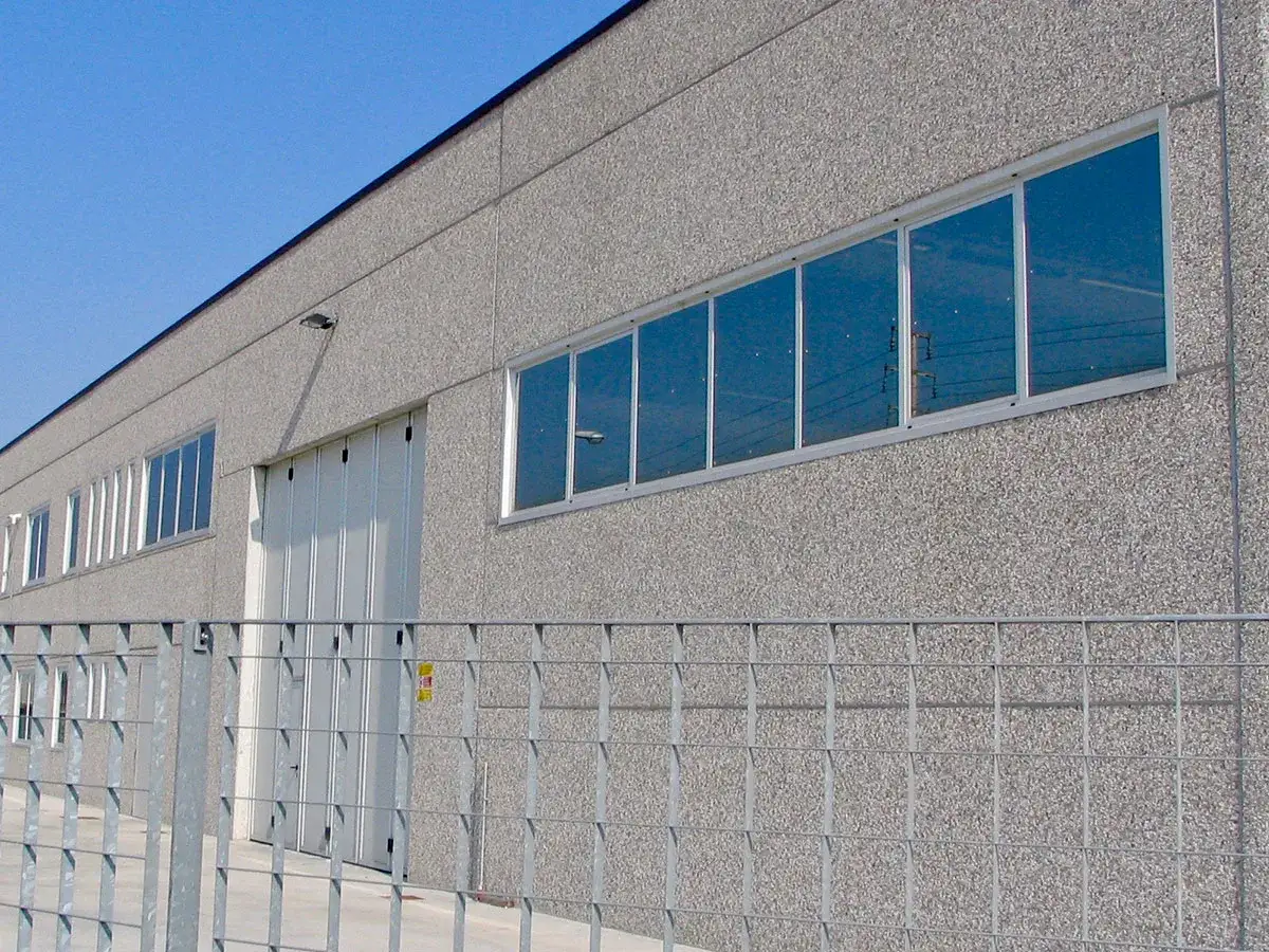Edificio industriale con pareti in cemento grezzo, una grande porta bianca a soffietto e finestre con infissi industriali che riflettono il cielo azzurro.