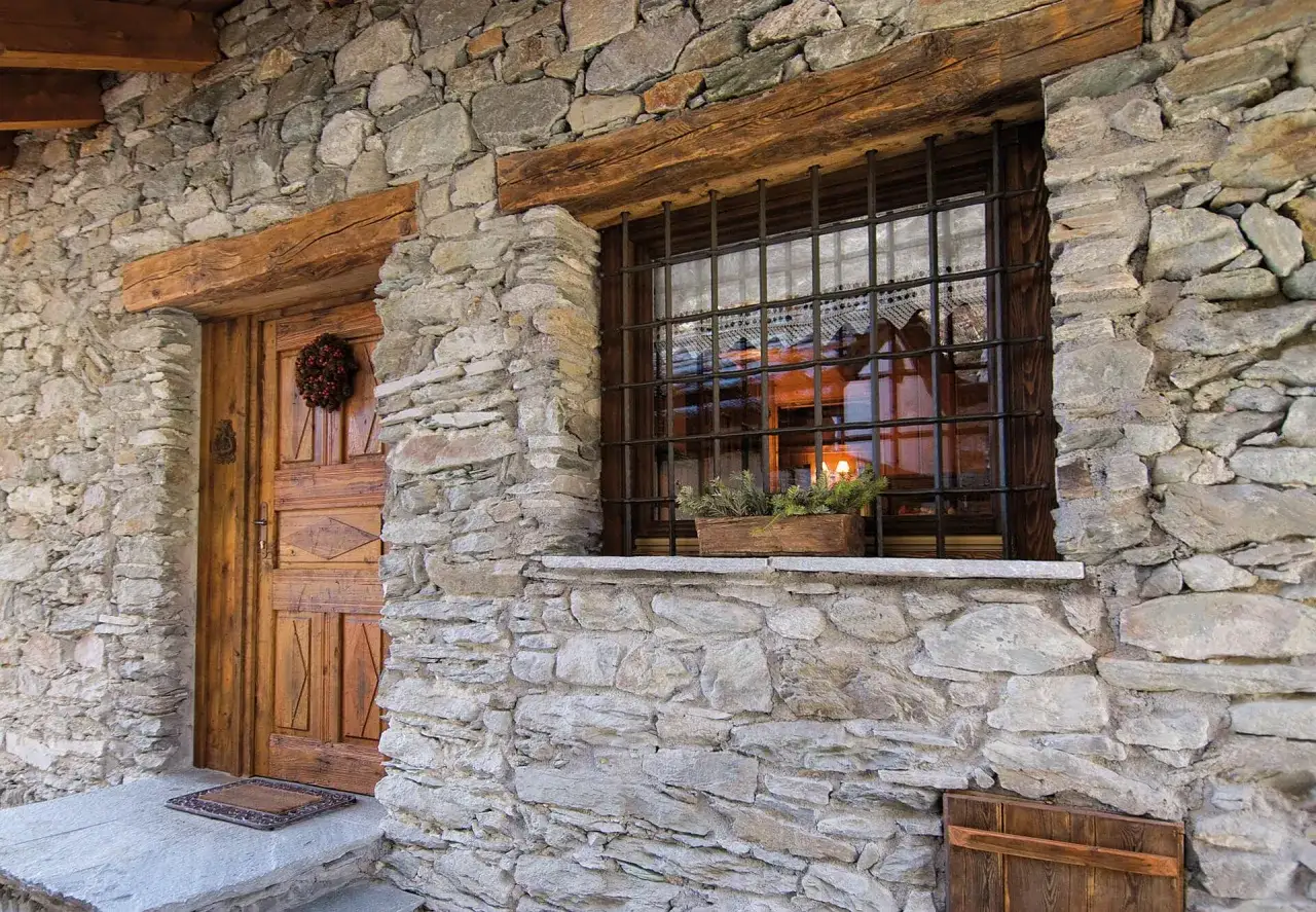 Muro in pietra di baita di montagna con porta in legno e finestre con inferriate.