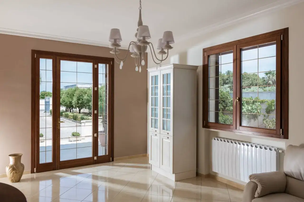 Serramenti legno alluminio pro e contro: un soggiorno luminoso con porte finestre e finestre in legno scuro, un armadio bianco e un radiatore moderno.