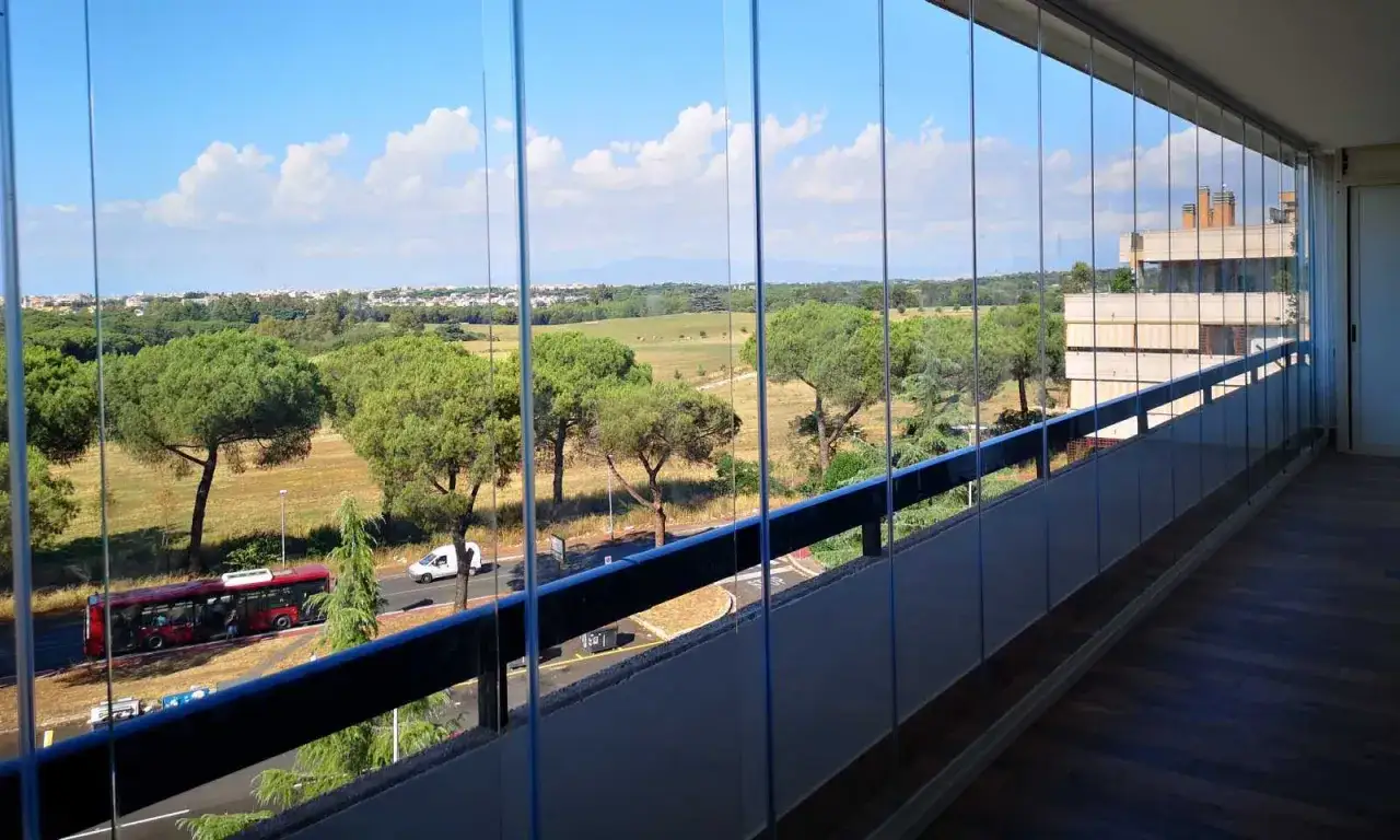 Vista dal balcone con vetrata condominio. Alberi, strada con autobus e un edificio sullo sfondo.