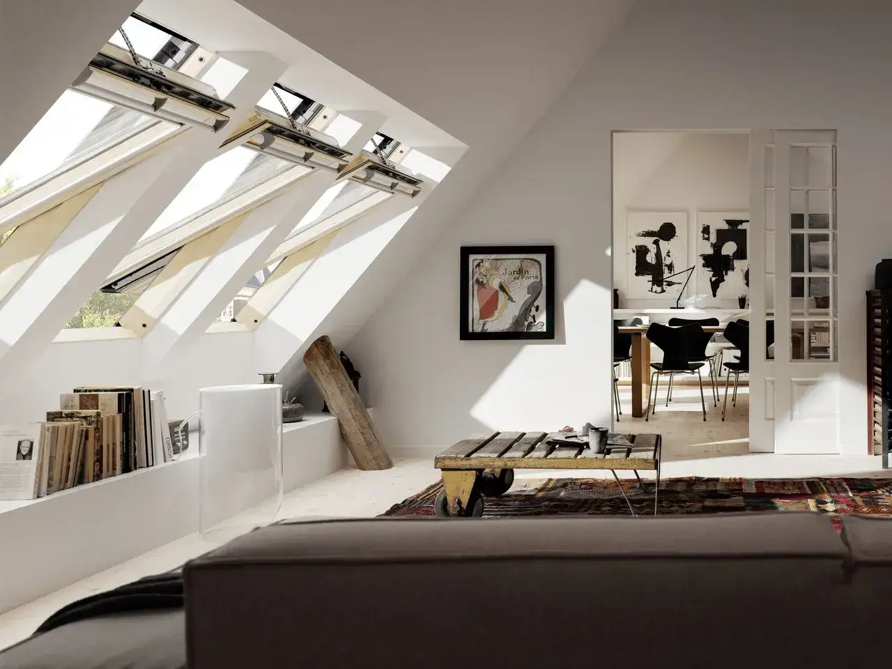 Luce naturale in mansarda grazie a finestre con apertura a bilico. Arredamento moderno con quadri astratti e un tavolino rustico.