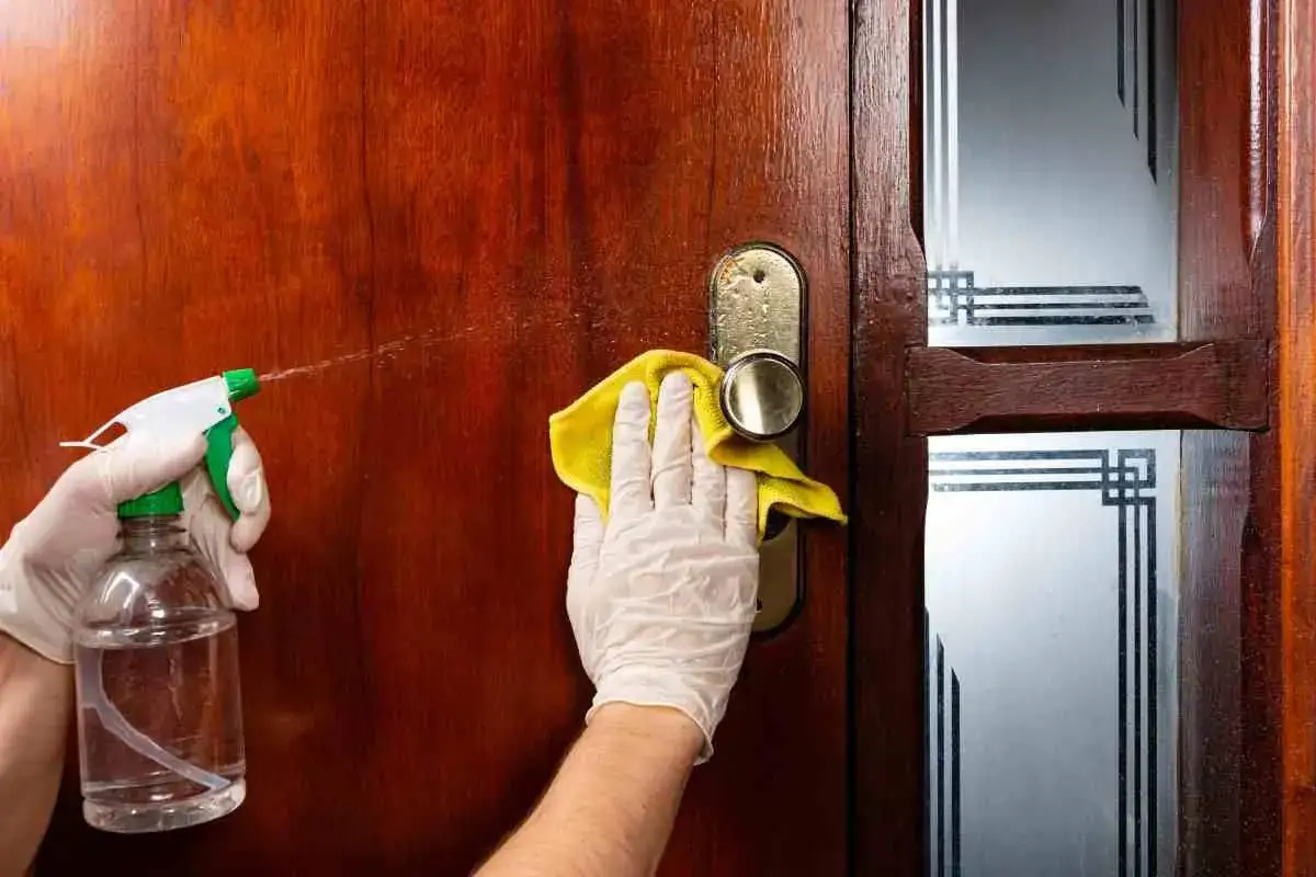 Mani guantate spruzzano detergente su una porta di legno scuro e la puliscono con un panno giallo.