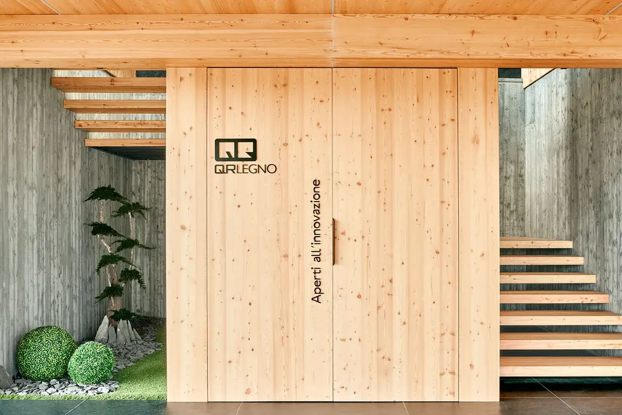 Porta scorrevole in infissi in legno lamellare con logo QR Legno. Scale in legno e cemento grezzo.